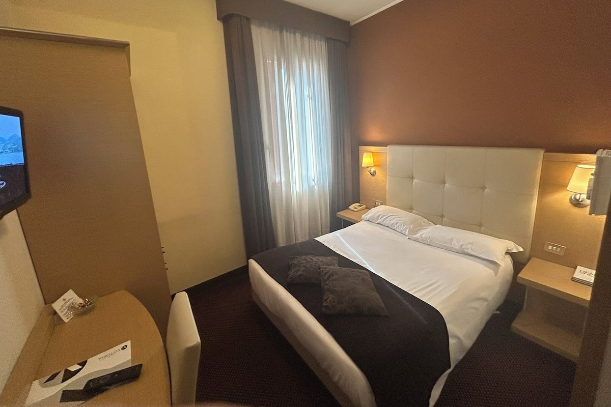 Una habitación de hotel con cama, televisión, escritorio y ventana con cortinas.
