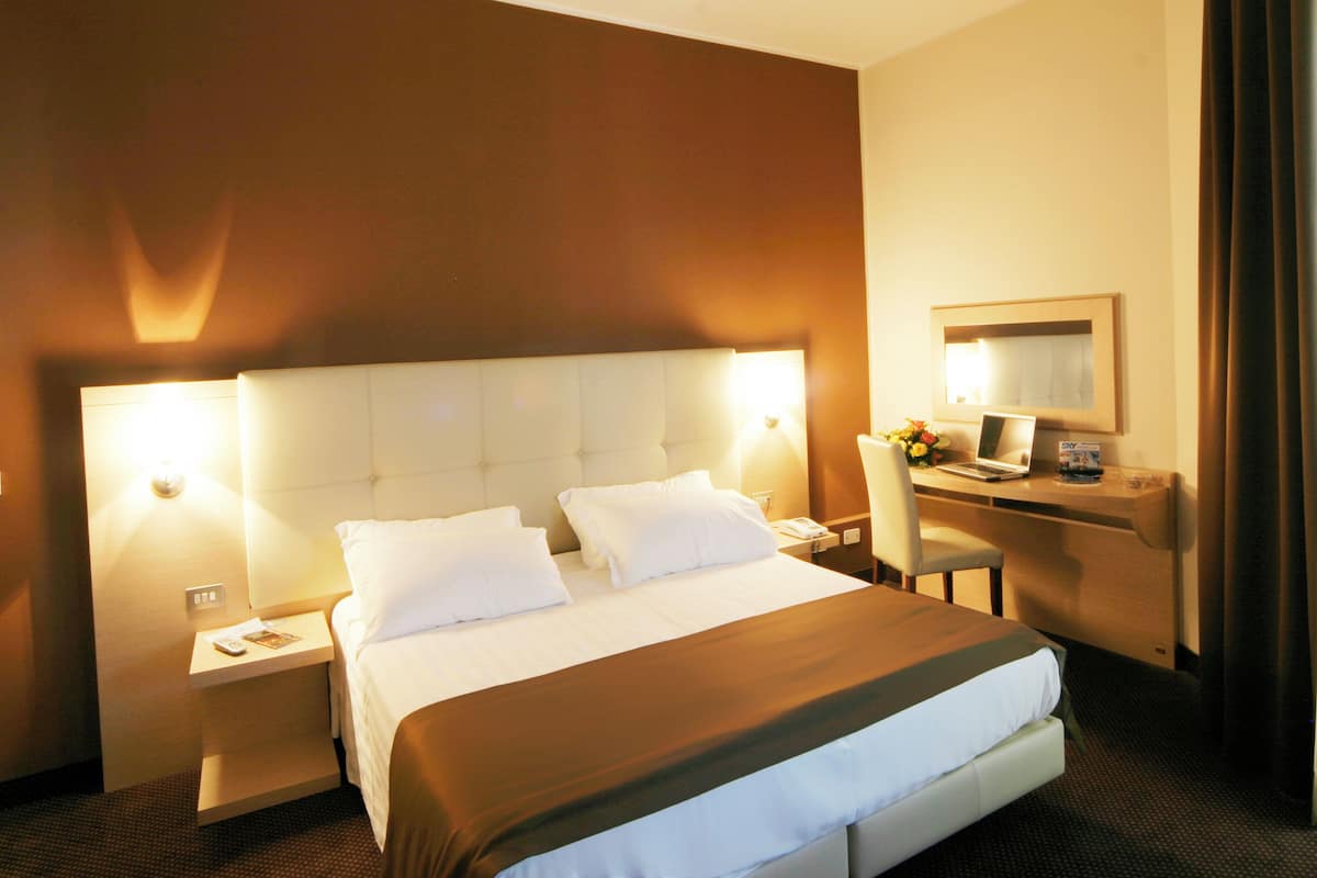 Habitación de hotel con una cama grande, un escritorio y una silla. Se observa un mesita de noche con una lámpara y un teléfono.