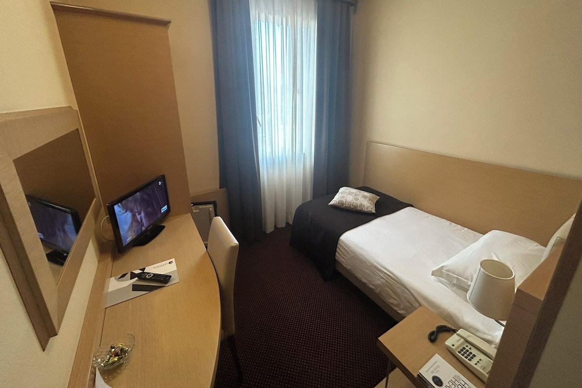 Una habitación de hotel con cama, escritorio con televisor, teléfono y ventana con cortinas.