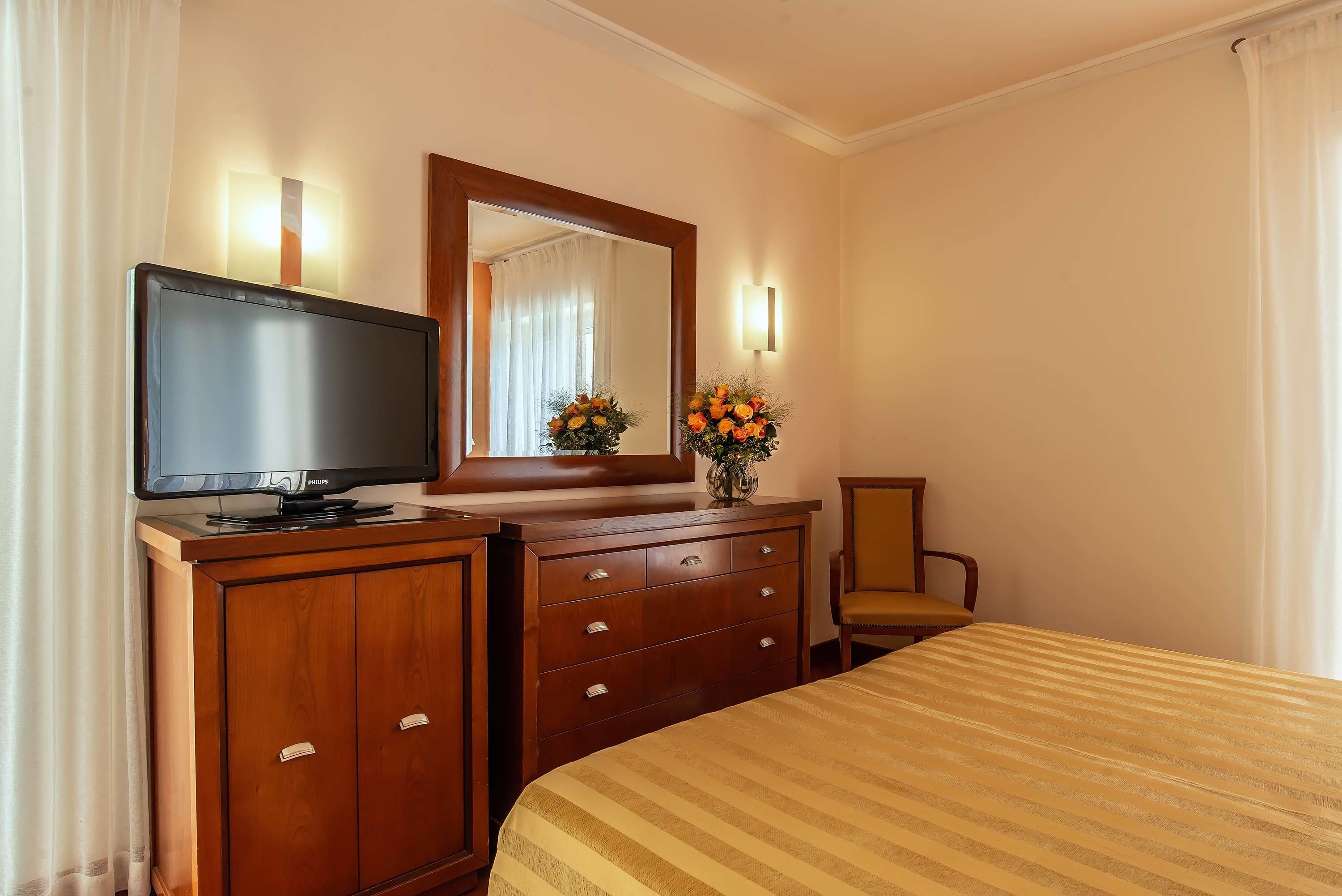 Une chambre d’hôtel avec un lit, un téléviseur posé sur une commode en bois, un miroir, un vase avec des fleurs et une chaise.