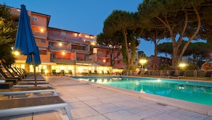 A swimming pool with lounge chairs and umbrellas, surrounded by a building with multiple balconies and trees. - Versilia Palace Hotel (Pietrasanta)