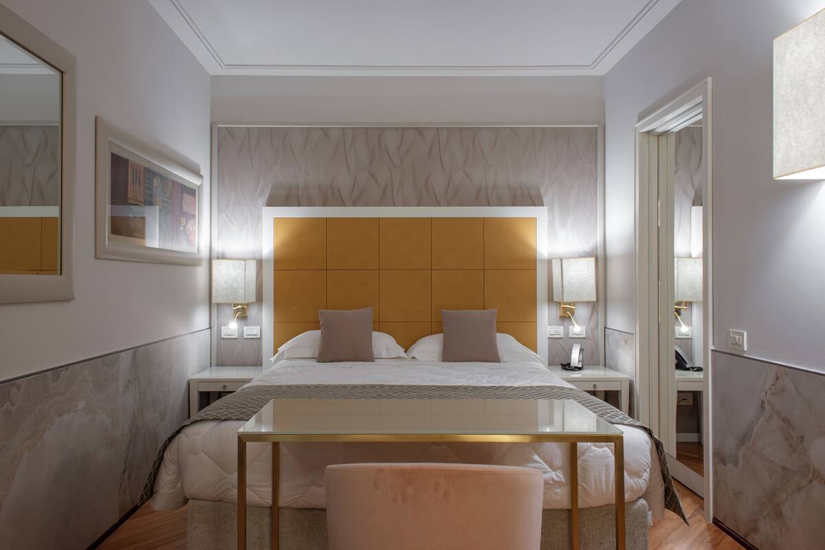 A modern hotel room with a large bed, bedside lamps, a nightstand, and a mirror.
