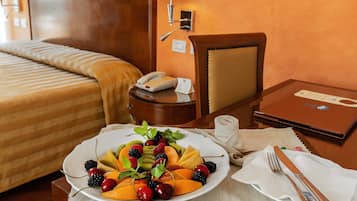 A hotel room with a bed, a bedside table with a lamp, a small table with a fruit platter, and a laptop on a desk.