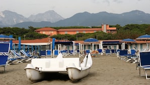 A beach with rows of blue lounge chairs and umbrellas, a white boat in the sand, and a building with red roofs in the background. - Versilia Palace Hotel (Pietrasanta)