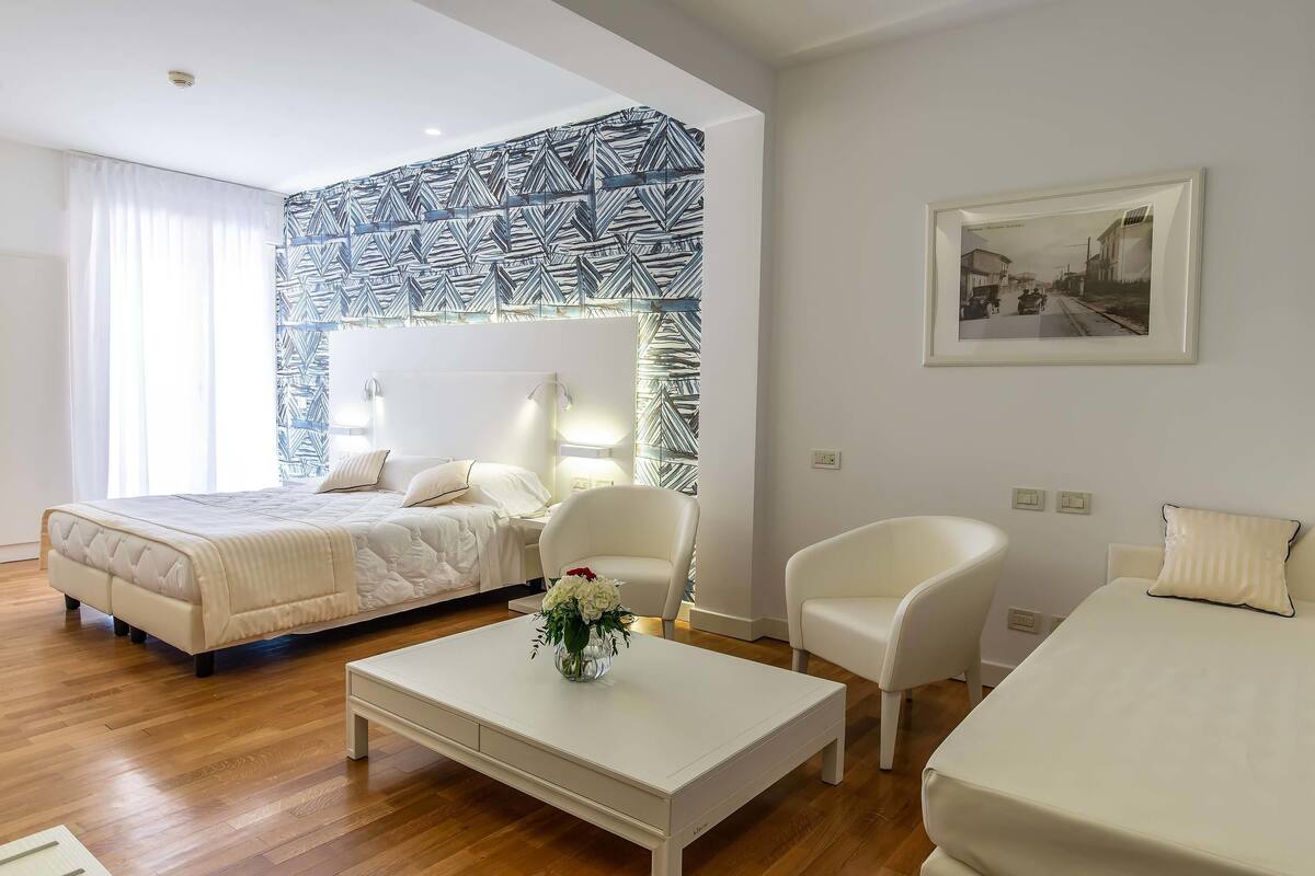 A modern hotel room with a bed, two white chairs, a coffee table with a vase, a patterned wall, and a framed picture.