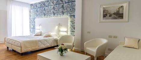 Una habitación de hotel moderna con una cama, dos sillas blancas, una mesa de centro con un florero, un papel tapiz estampado y un cuadro enmarcado.