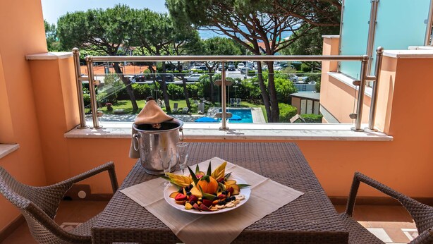 A balcony with wicker furniture, a table with a fruit platter, a metal bucket, and a view of a swimming pool and trees. - Versilia Palace Hotel (Pietrasanta)