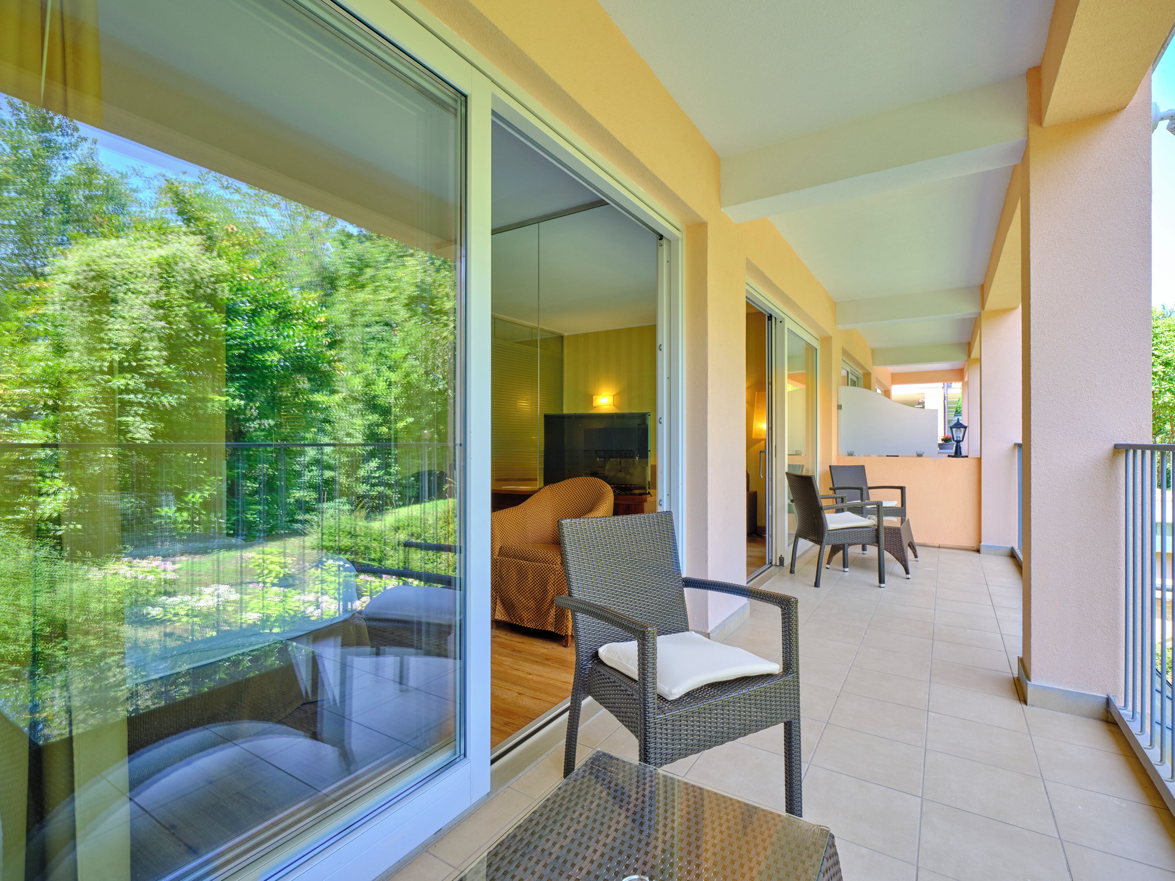 A balcony with a glass door leading to a living area with a sofa and chairs, a TV, and a view of greenery.