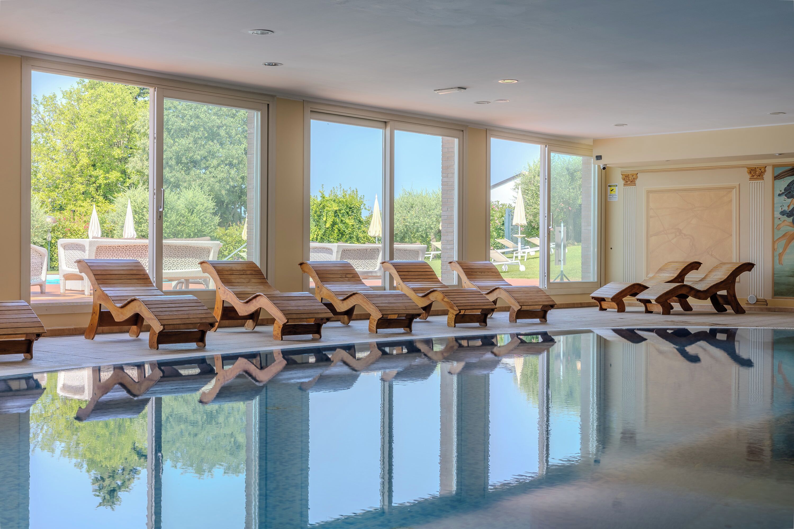 A swimming pool with wooden loungers and large windows offering a view of trees and a patio area.