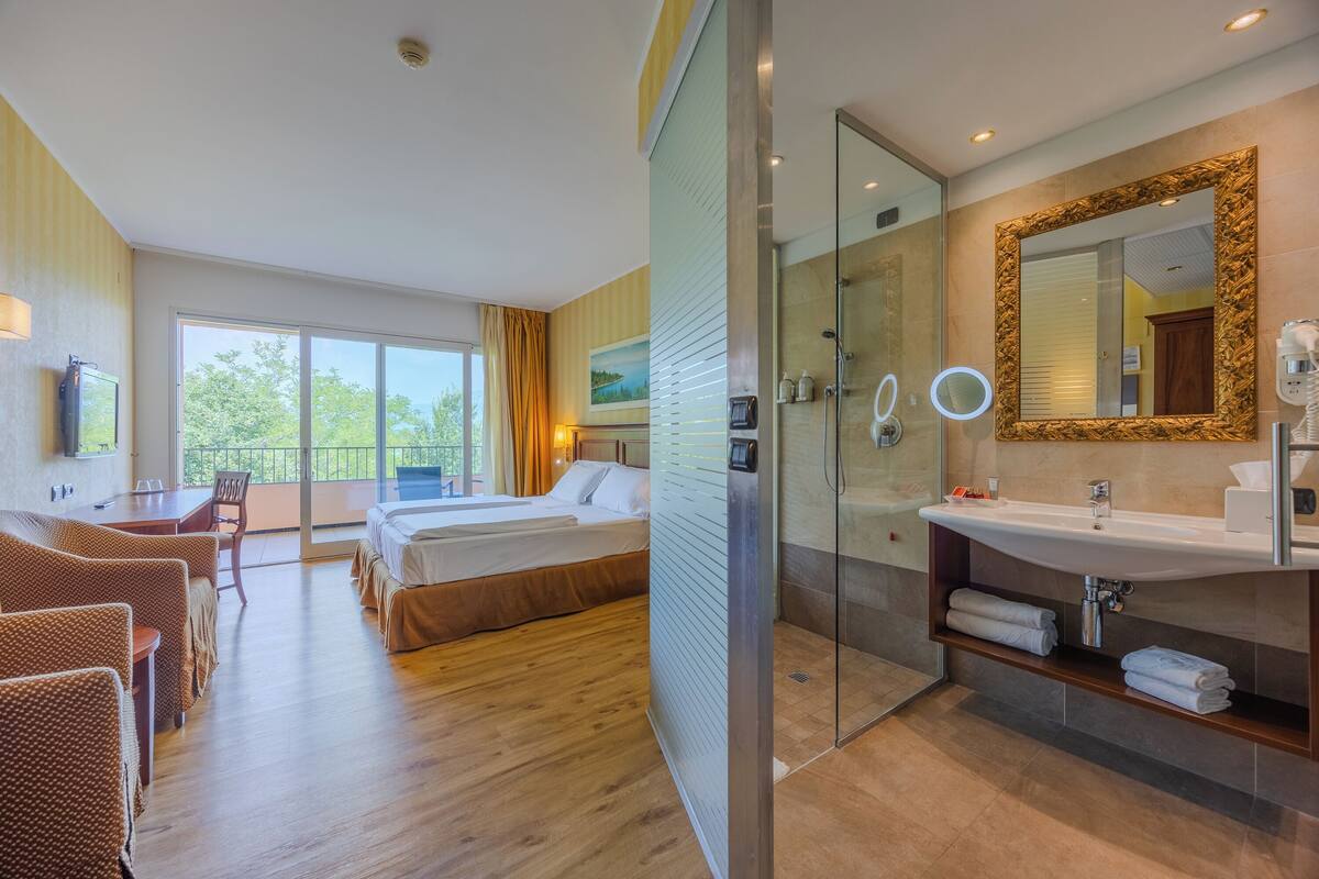 A hotel room with a large bed, a separate seating area, a bathroom with a mirror and sink, and a view of greenery outside.