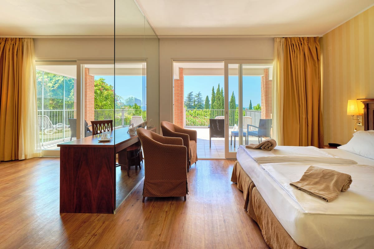 A hotel room with a wooden floor, a bed with a beige bedspread, a desk with a chair, and a balcony with chairs.