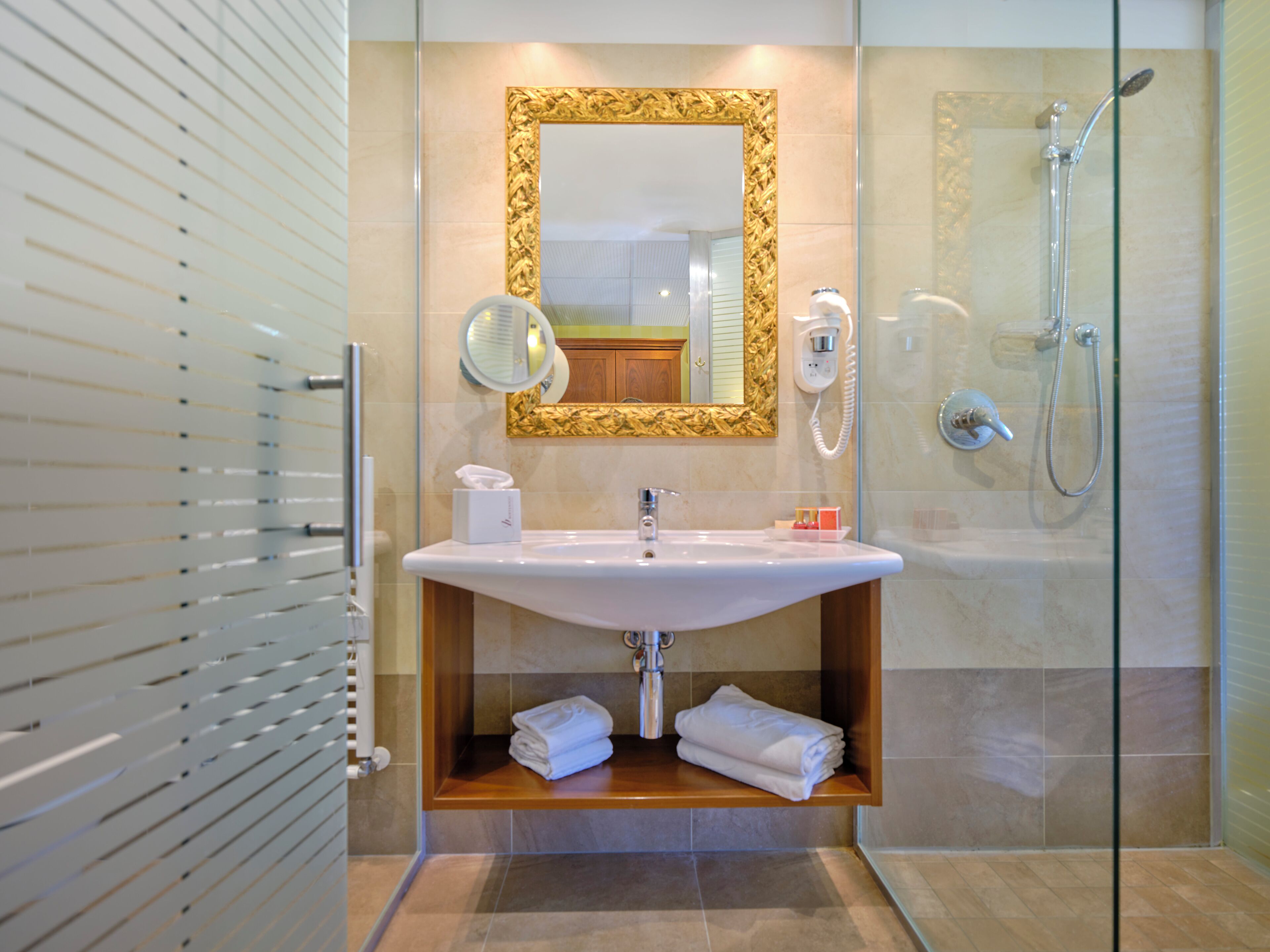 A modern bathroom with a glass shower enclosure, a white sink with a gold-framed mirror, and neatly folded towels on a wooden shelf.