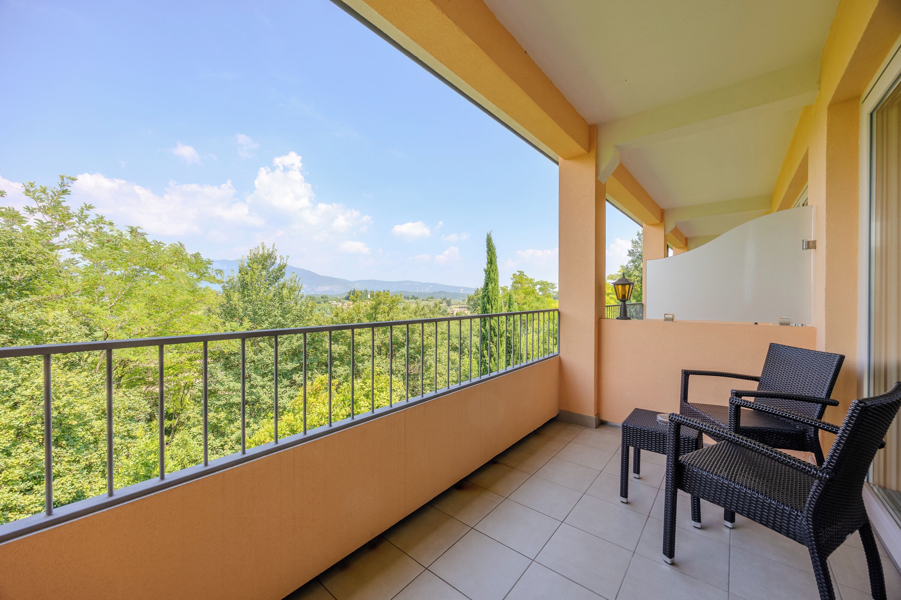 A balcony with a view of greenery and mountains, a tiled floor, and two wicker chairs.