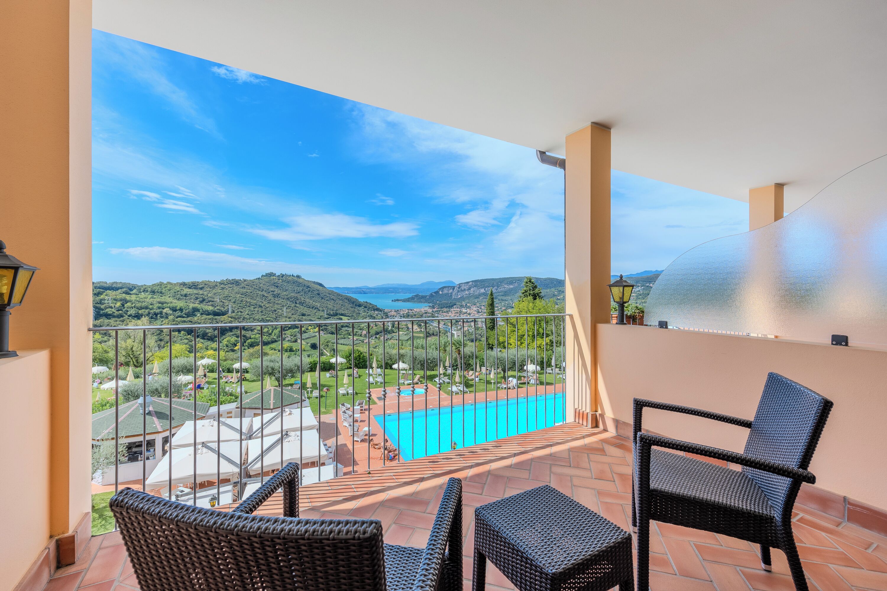 Un balcon avec des fauteuils en osier, une vue sur une piscine et des collines lointaines.