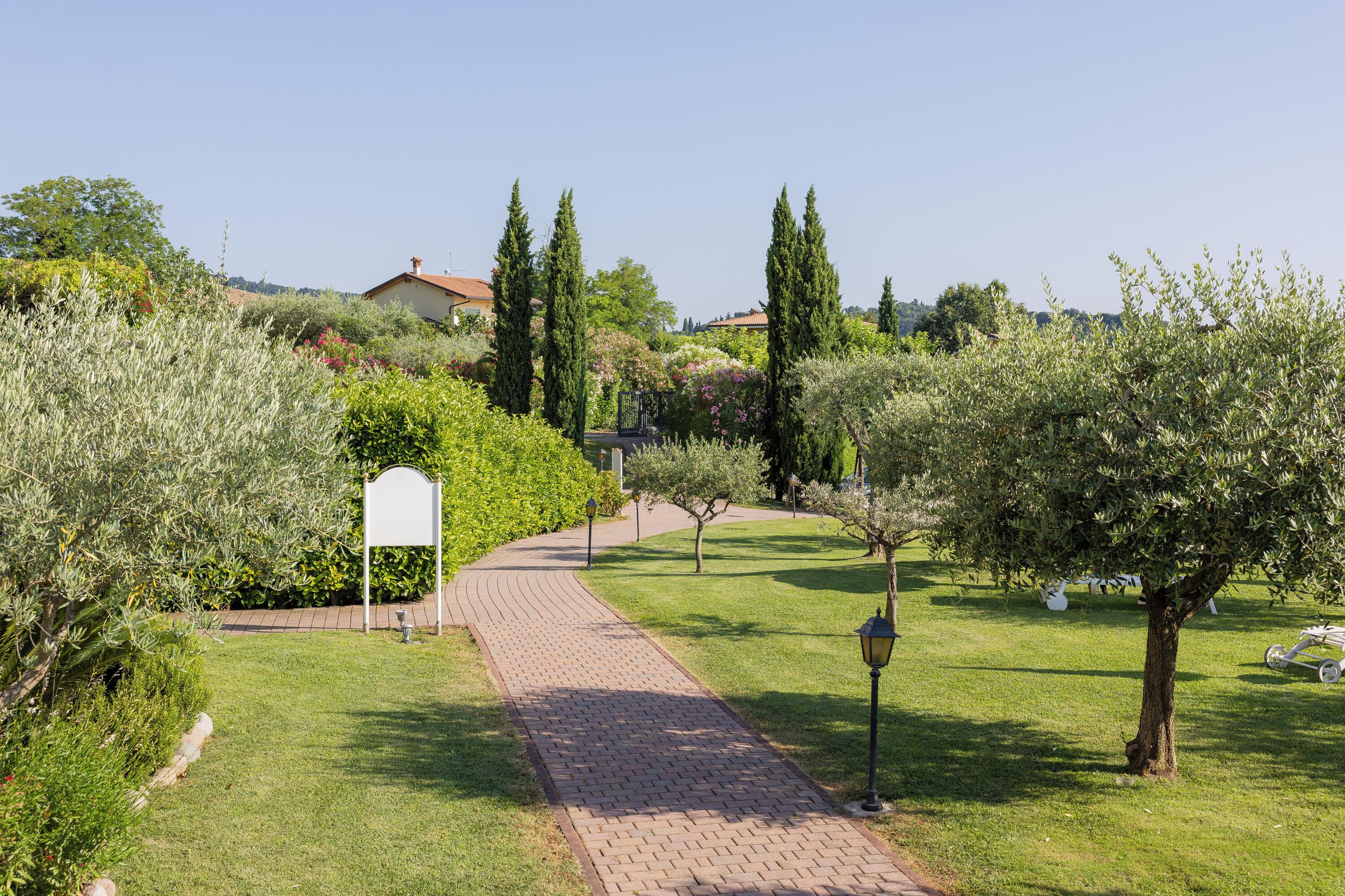 Un chemin pavé traverse un jardin bien entretenu, où l’on trouve différentes espèces d’arbres et d’arbustes.