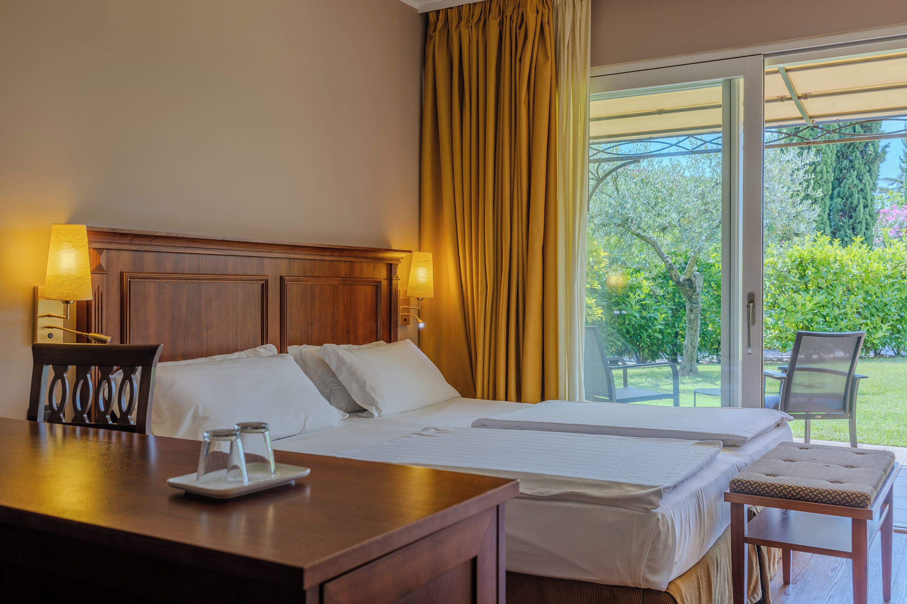 Habitación de hotel con una cama grande, una mesita de noche de madera con una lámpara, una pequeña mesa con dos vasos y vista a un jardín por una puerta corrediza de vidrio.