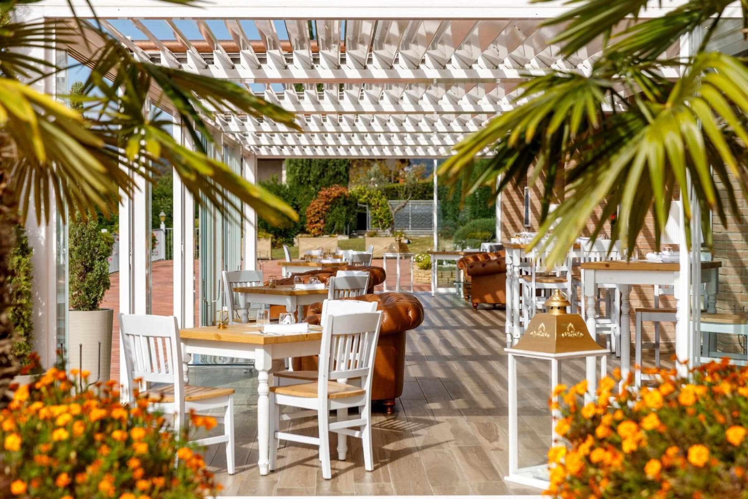 An outdoor dining area with white tables and chairs, a pergola overhead, and potted plants.