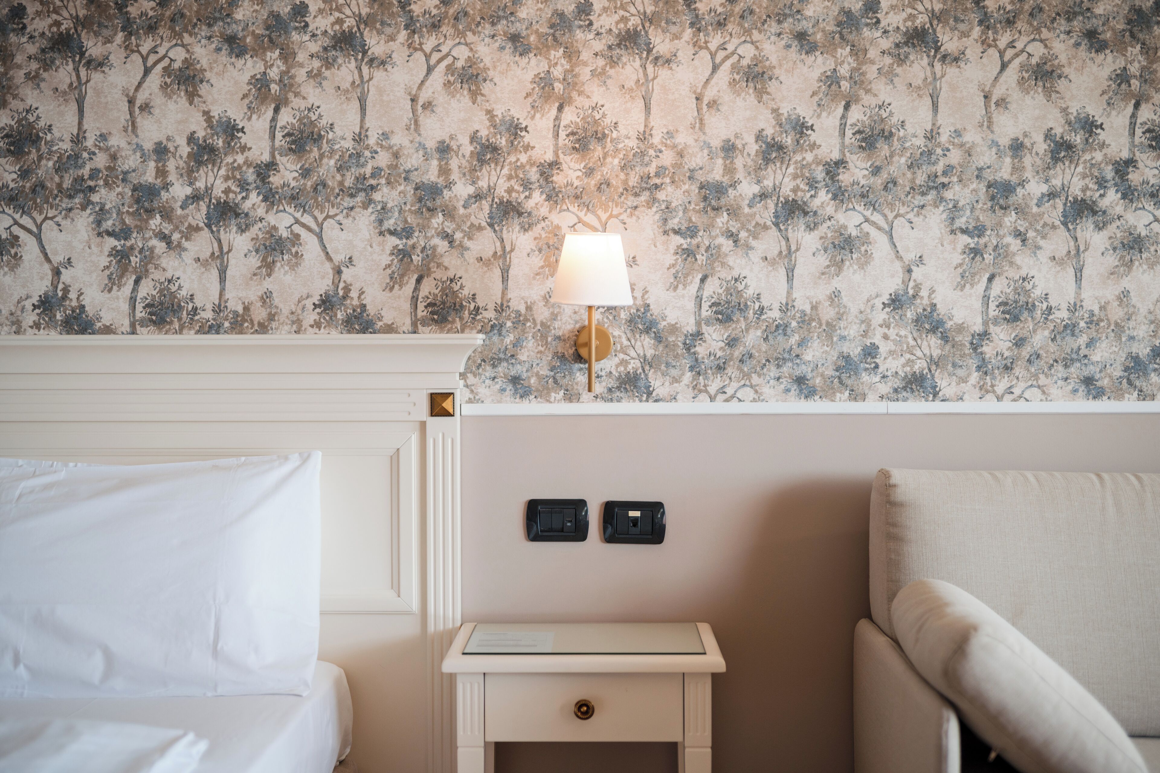 A hotel room with a floral wallpaper, a bedside lamp, a nightstand with two switches, and a bed with white bedding.