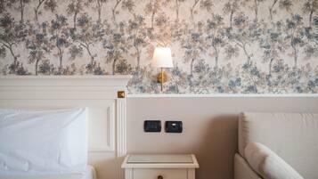 Una habitación de hotel con papel tapiz floral, una lámpara de noche, una mesita de noche con dos interruptores y una cama con ropa blanca.