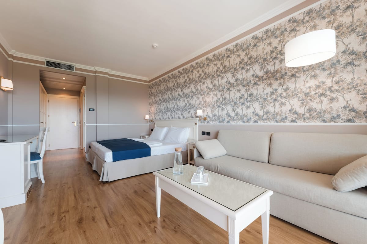 A modern hotel room with a bed, a sofa, a glass-top table, and a wall with floral wallpaper.
