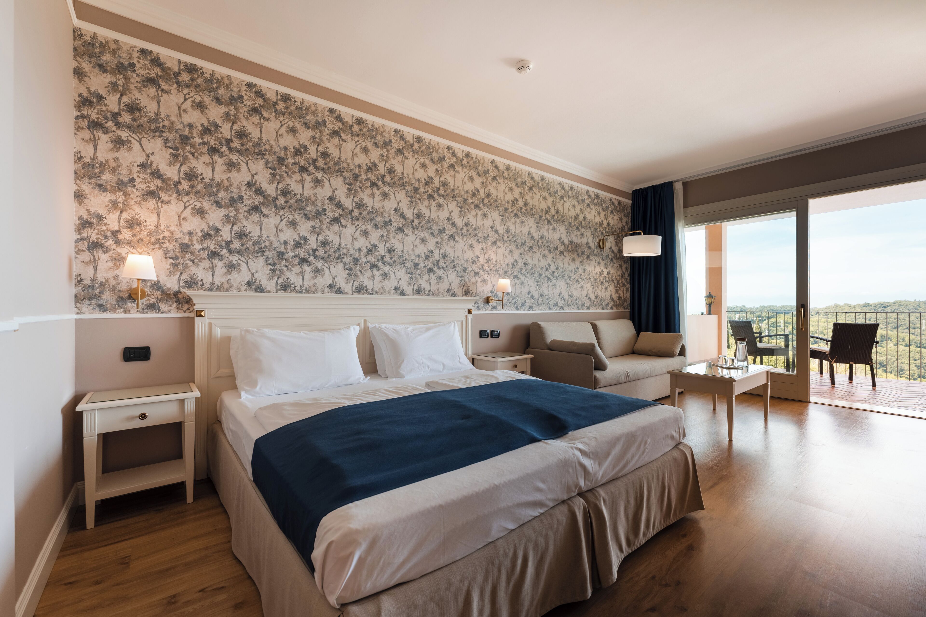 Una habitación de hotel con una cama grande, un sofá, un balcón con vistas y una mesita de noche con una lámpara.