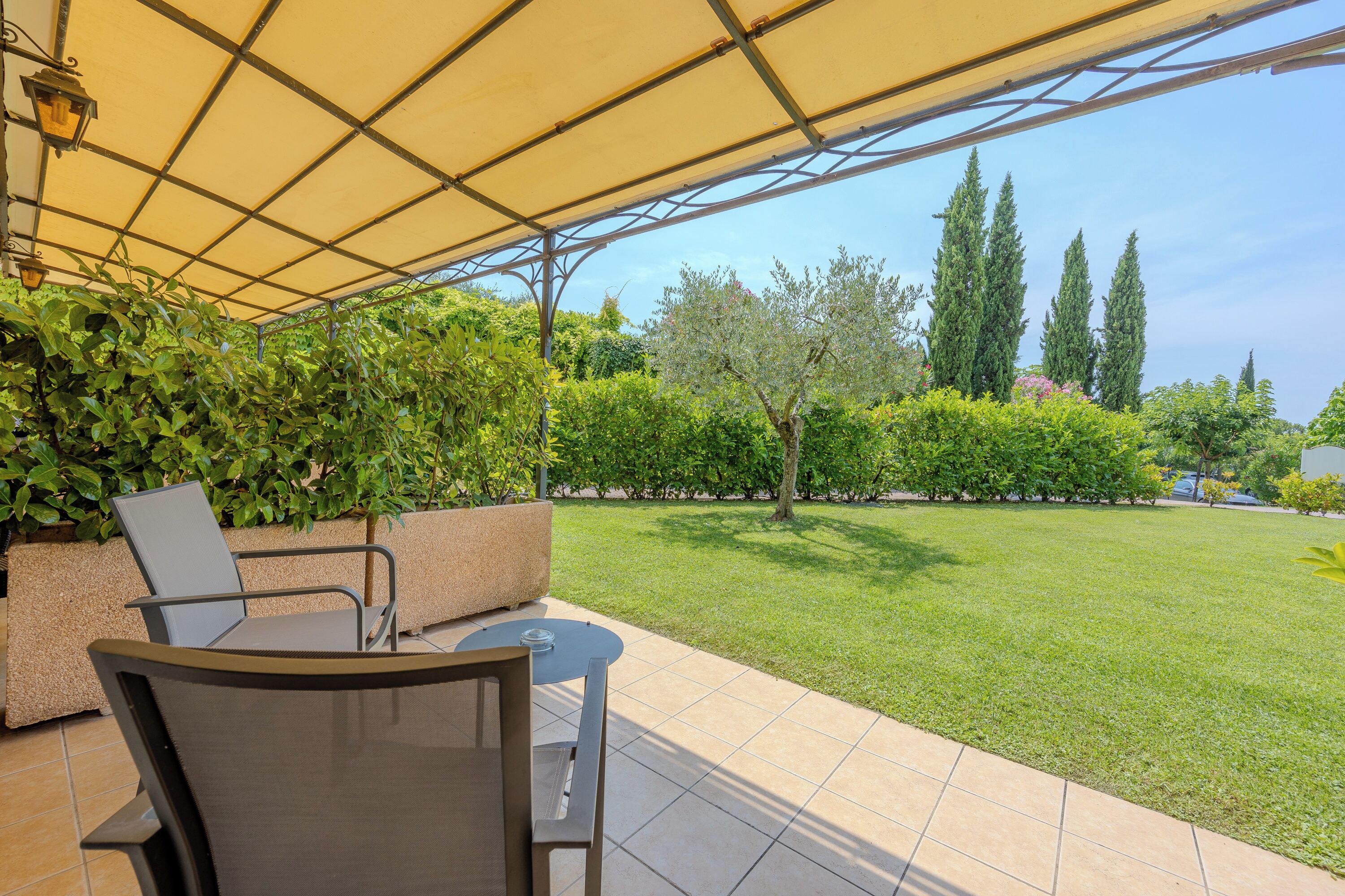 An outdoor patio area with a table and chairs, a yellow canopy, and a well-maintained lawn with trees and shrubs.
