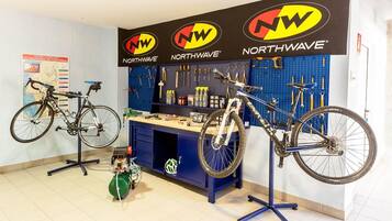 Un taller de reparación de bicicletas con dos bicicletas en caballetes, un banco de trabajo con herramientas y un tablero de anuncios con utensilios.