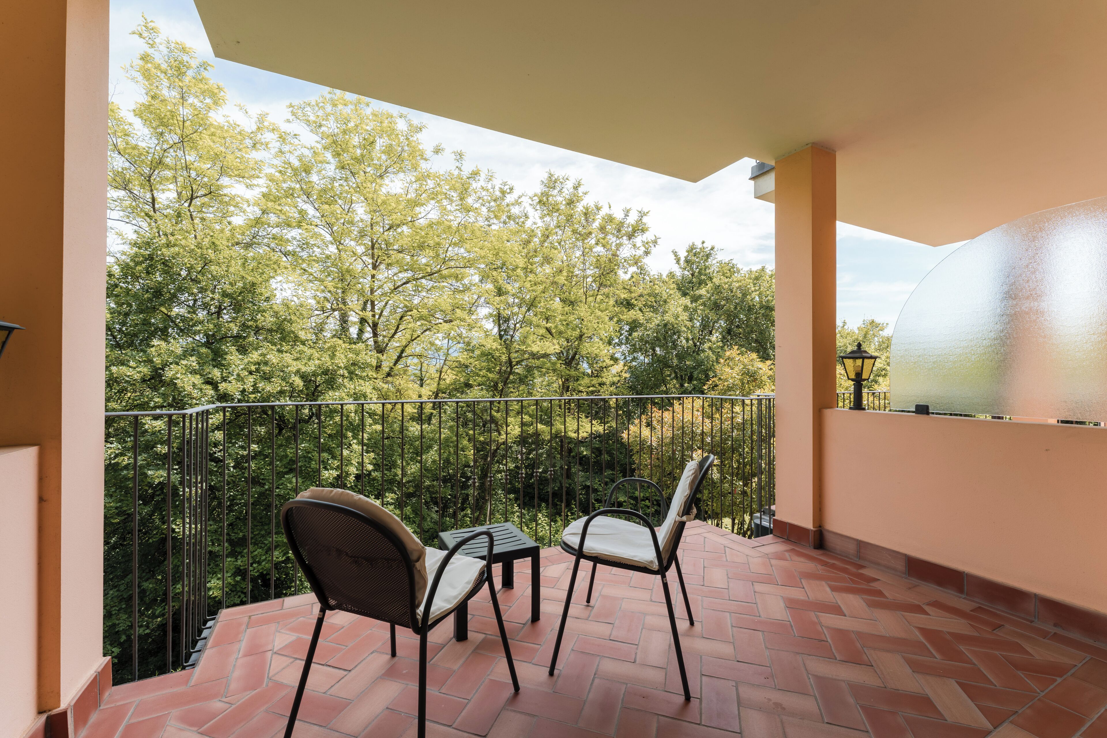 Un balcón con dos sillas y una mesa, rodeado por una barandilla de metal, con vistas a un jardín con árboles.