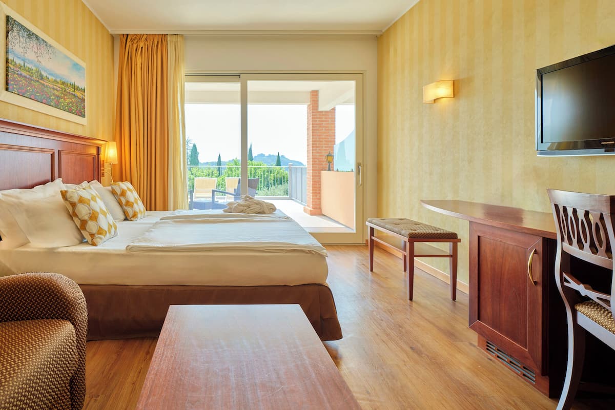 A hotel room with a bed, a desk, a chair, a television, and a balcony with a view of greenery.