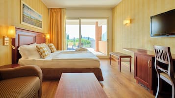 Habitación de hotel con cama, escritorio, silla, televisor y balcón con vistas a zonas verdes.