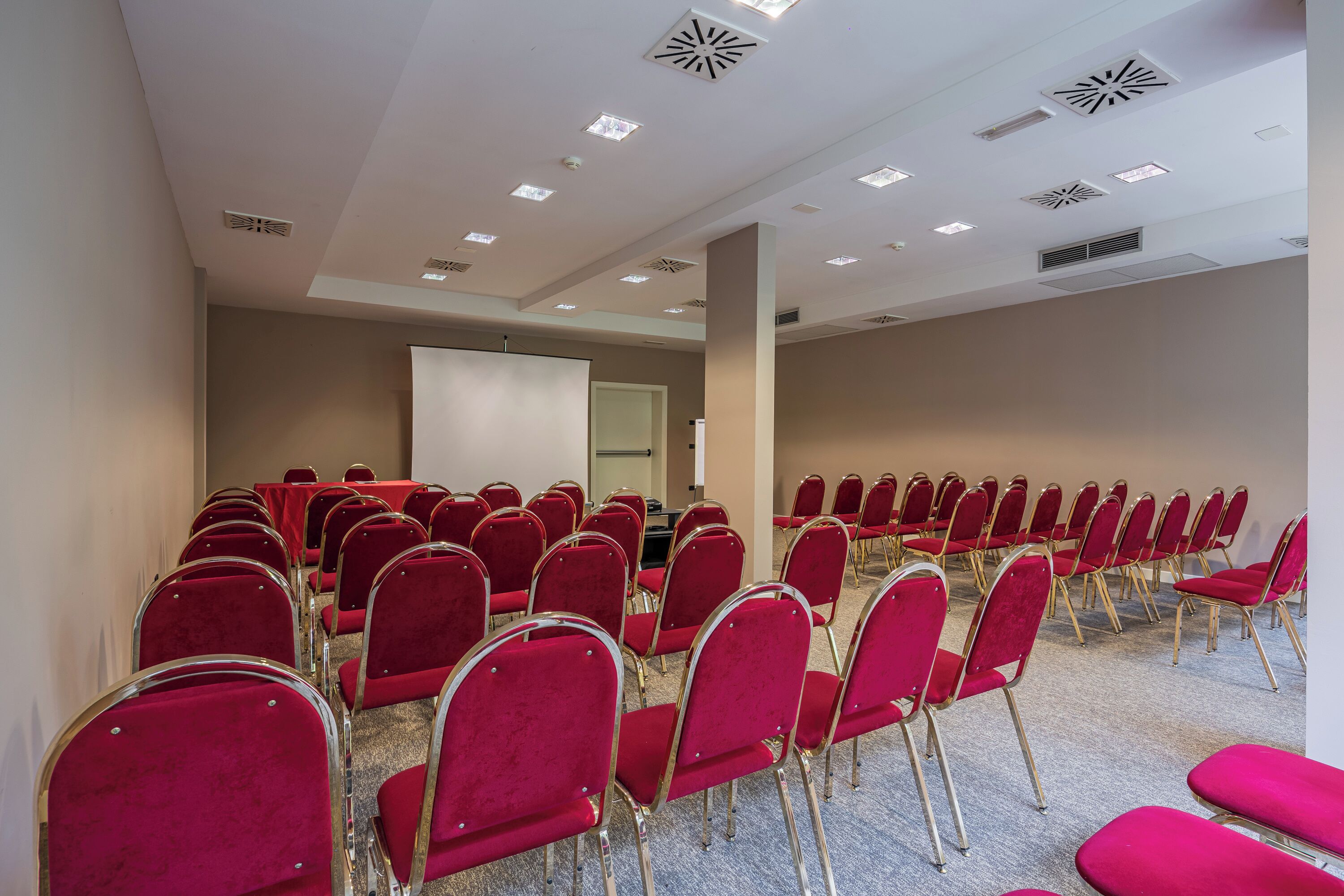 A conference room with rows of red chairs, a whiteboard, and a projector screen.