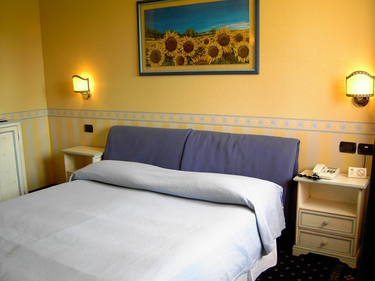 A hotel room with a bed, a nightstand with a telephone, and a wall-mounted lamp.