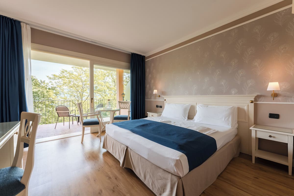 A hotel room with a large bed, a desk, a chair, and a balcony with a table and chairs.