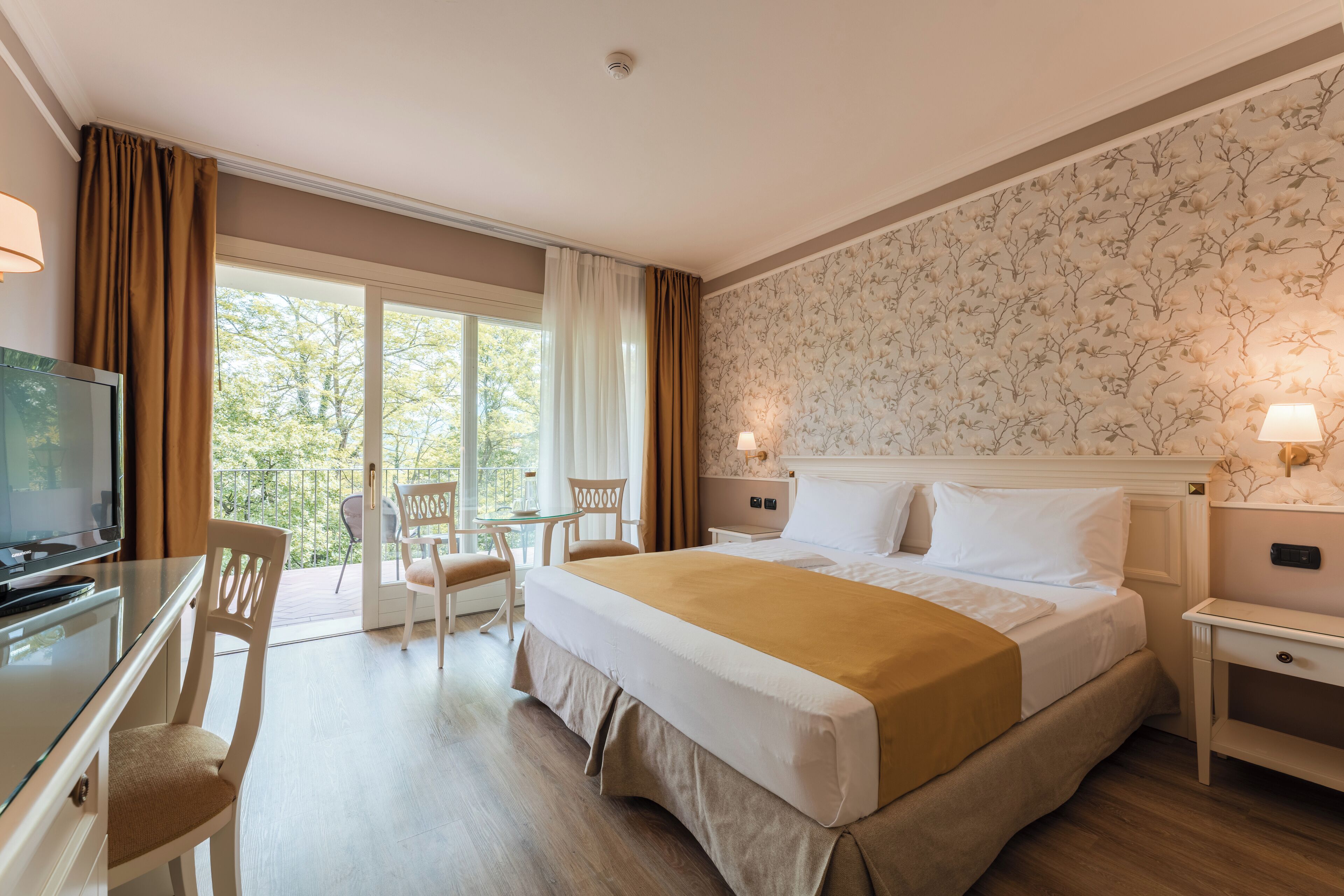 A hotel room with a bed, a desk, a TV, and a balcony with chairs.