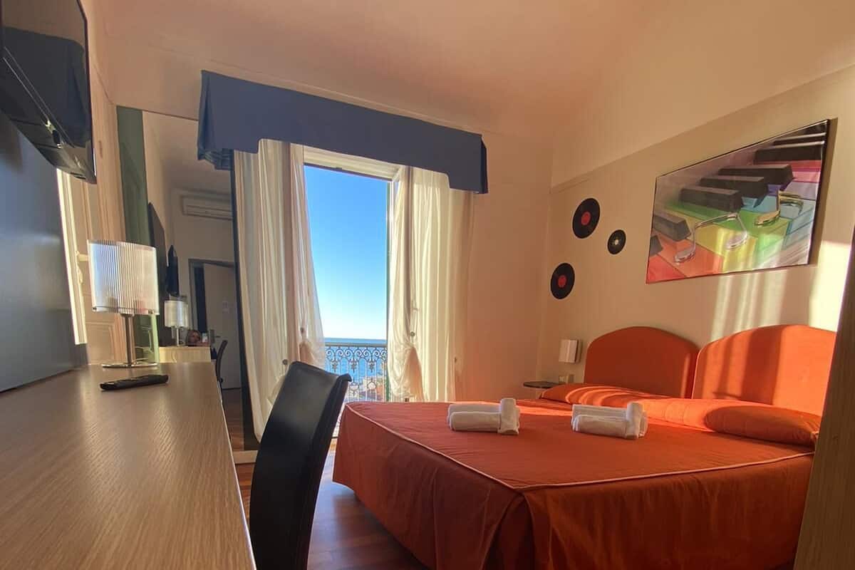 A hotel room with a wooden desk, a bed with orange bedding, a chair, and a view of the sky through the window.