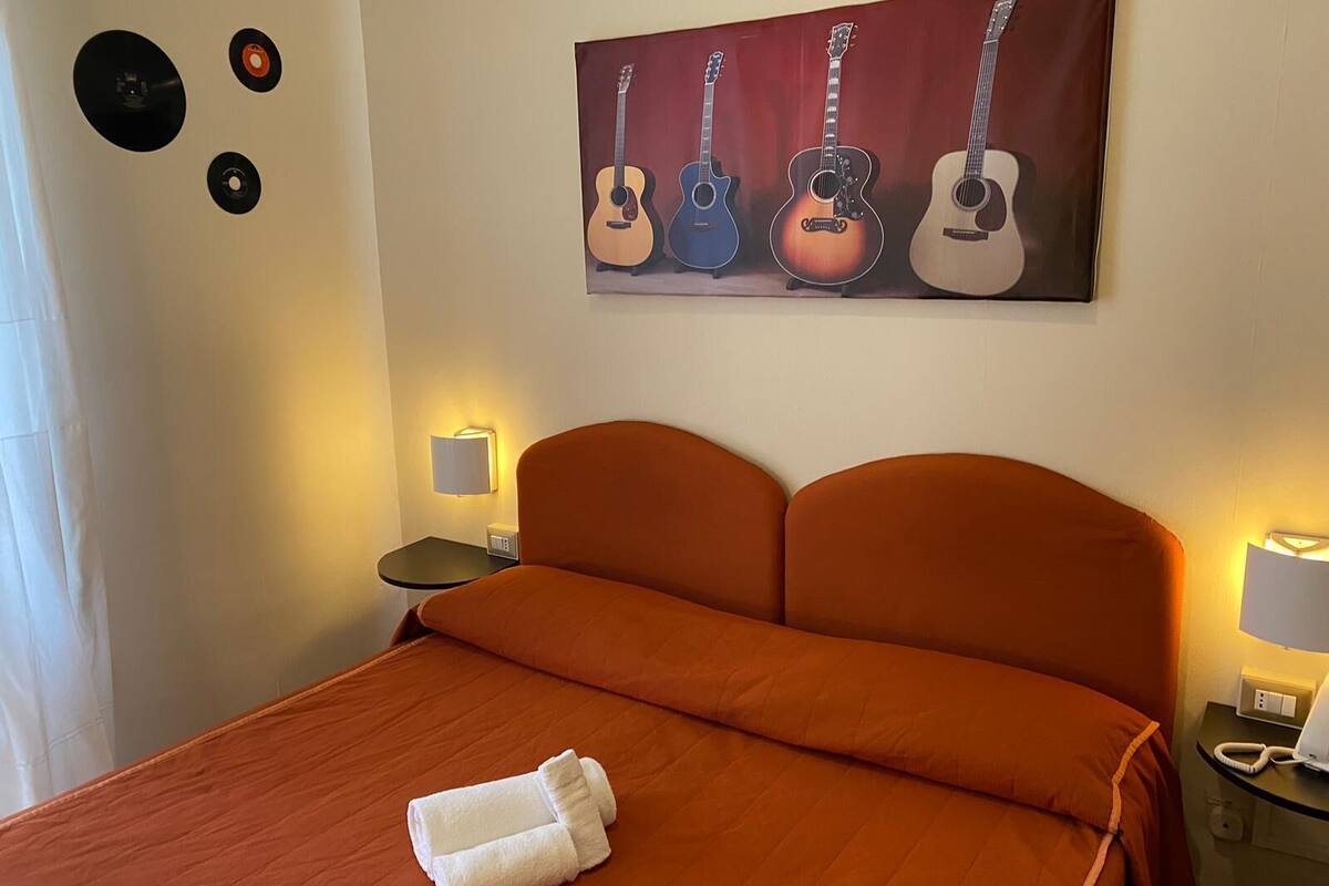 A hotel room with a bed, a painting of guitars, a lamp, and a telephone on the nightstand.