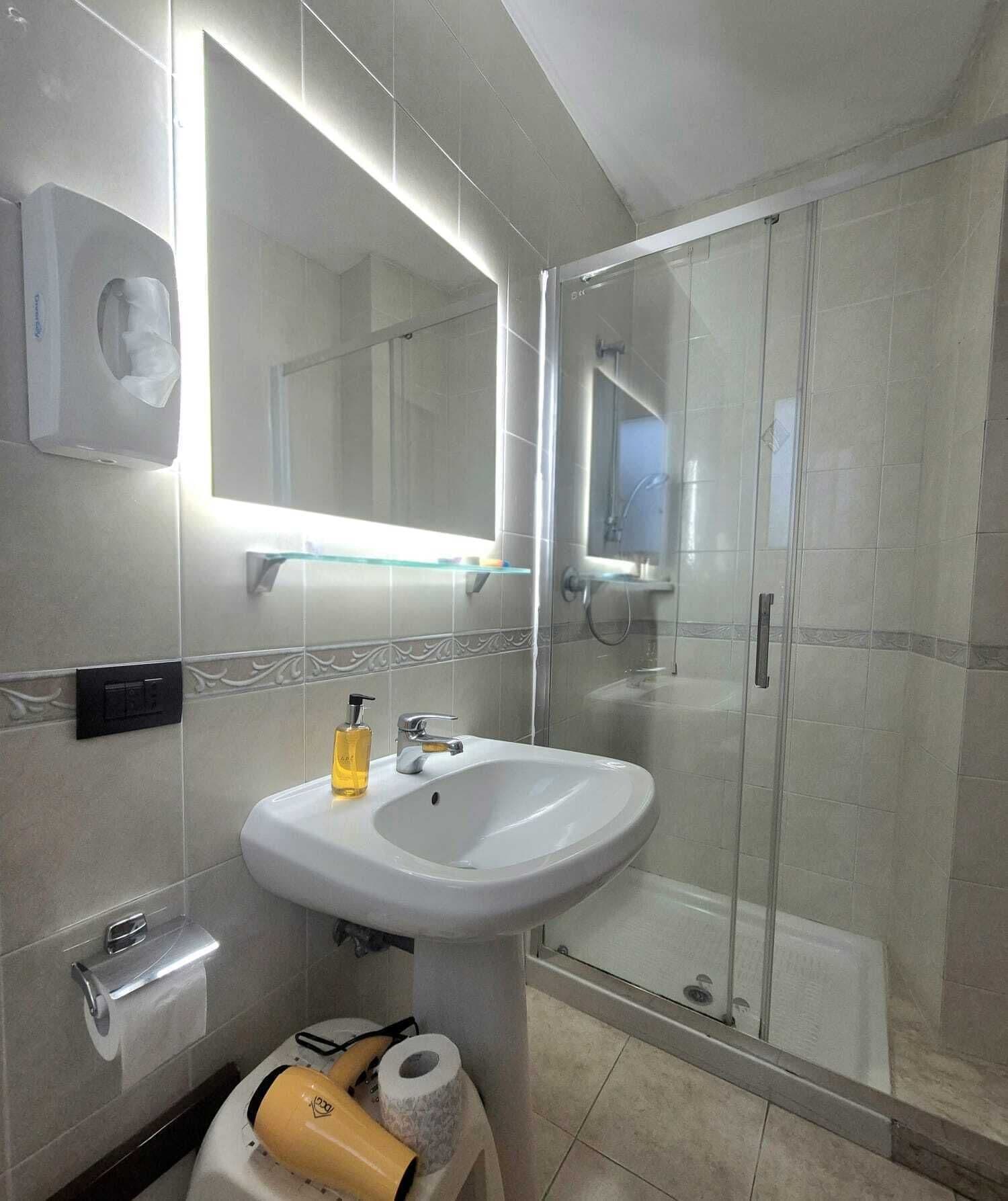 Un baño moderno con ducha envuelta en vidrio, un lavabo blanco con dispensador de jabón y un espejo con iluminación integrada.