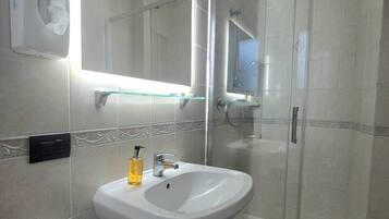 Un baño moderno con ducha envuelta en vidrio, un lavabo blanco con dispensador de jabón y un espejo con iluminación integrada.