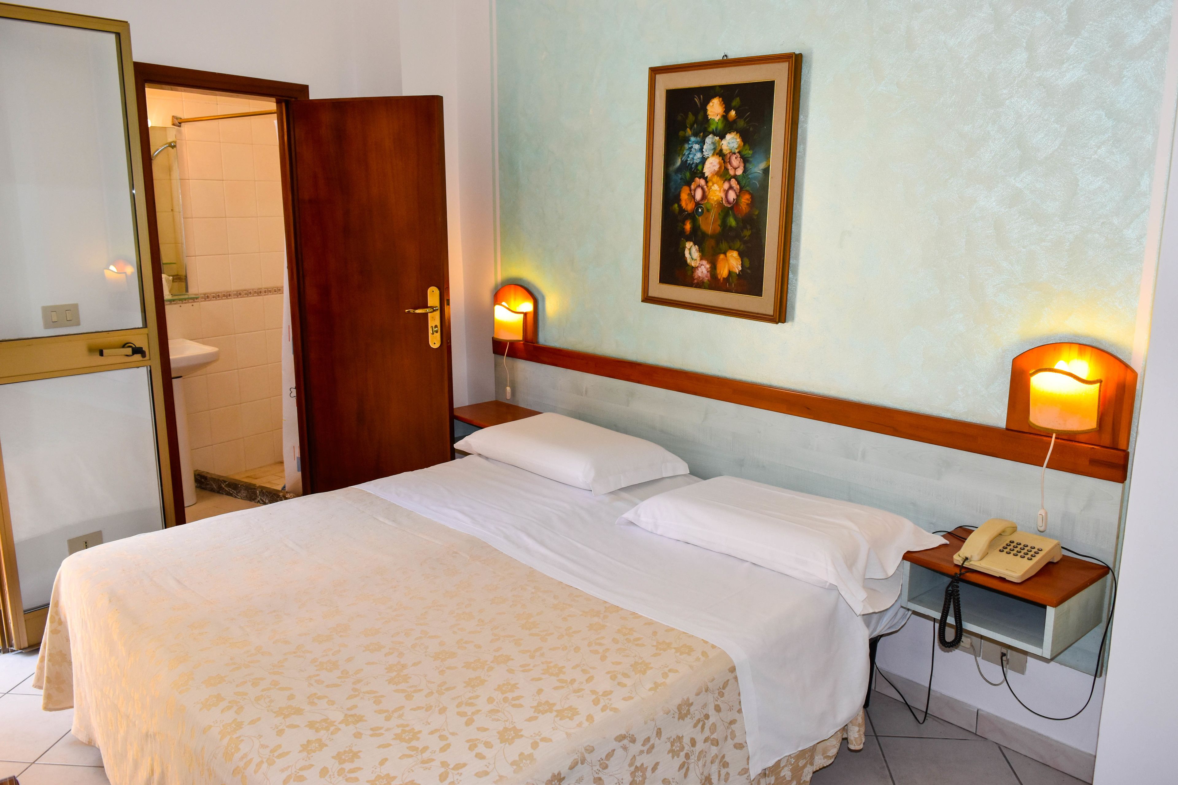 Una habitación de hotel con una cama, un teléfono en la mesita de noche, un cuadro en la pared y un baño visible a través de una puerta abierta.