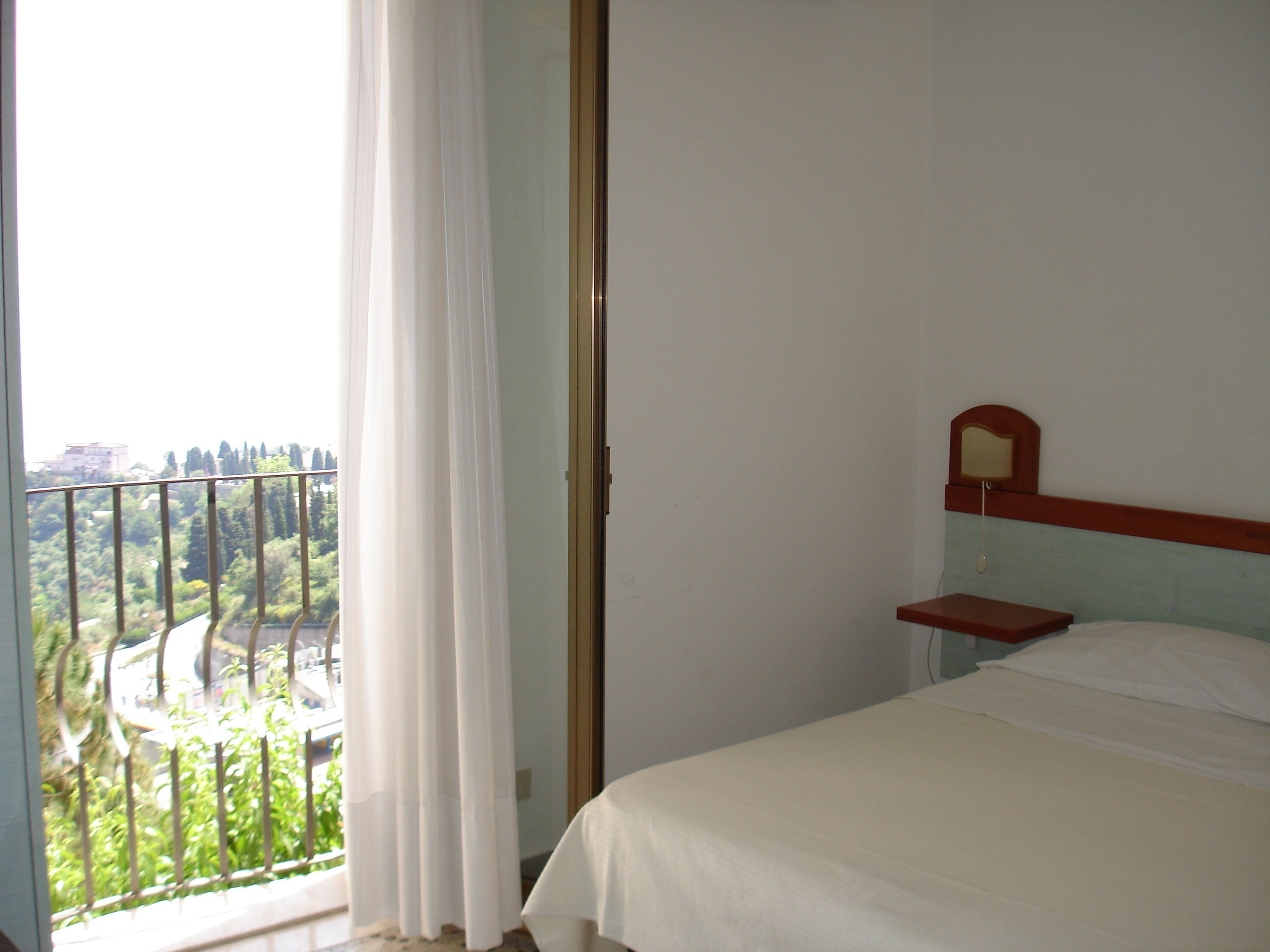 Un dormitorio con una cama, una mesita de noche y vistas a zonas verdes y edificios a través de una ventana con cortina.