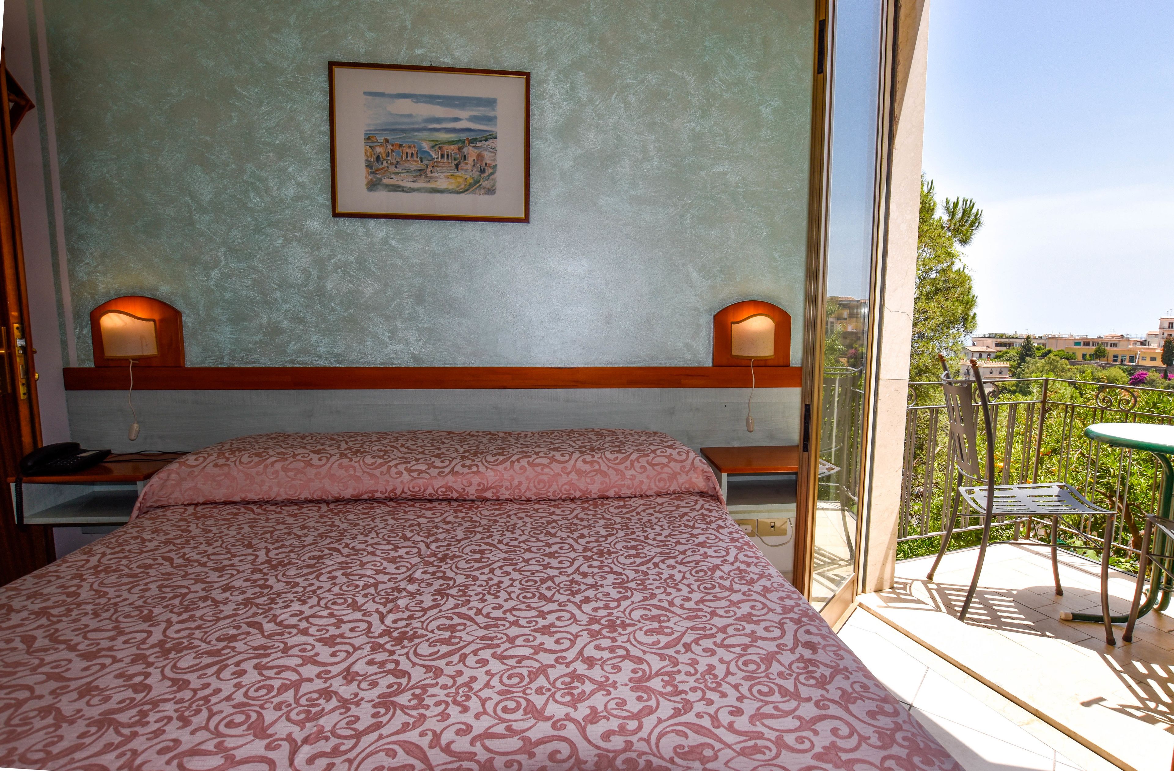 Ein Schlafzimmer mit einem gemusterten Bett, zwei Nachttischlampen, einem gerahmten Bild an der Wand und einem Balkon mit Tisch und Stühlen.