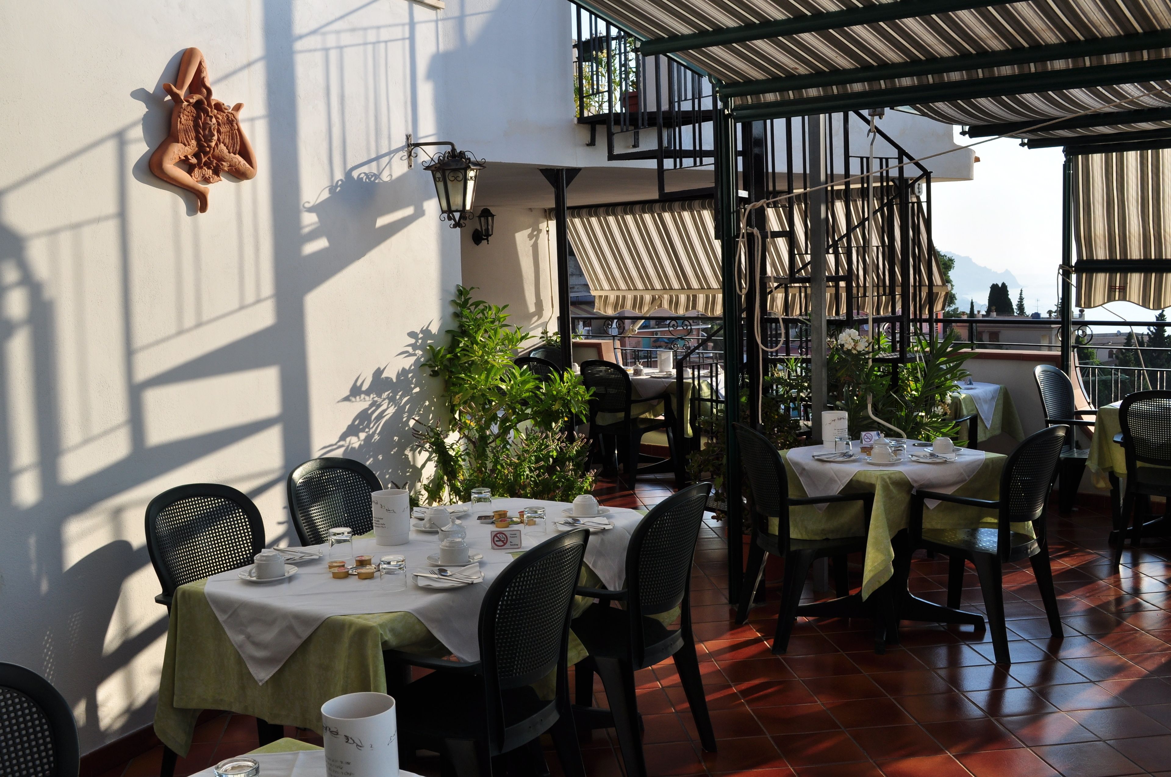 A restaurant with outdoor seating, tables set with white tablecloths, black chairs, and potted plants.
