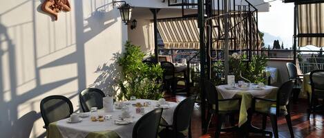 A restaurant with outdoor seating, tables set with white tablecloths, black chairs, and potted plants.