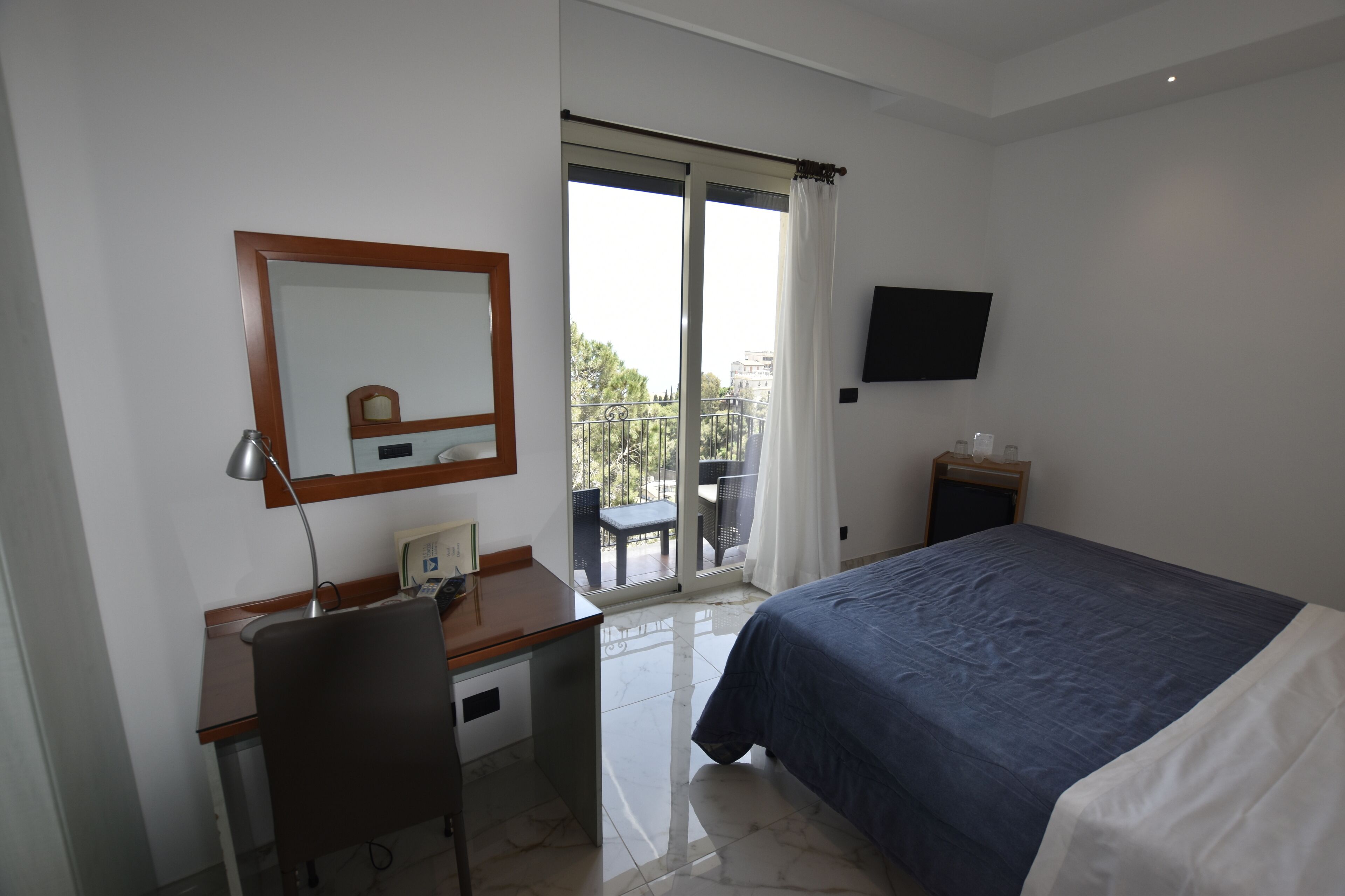 A hotel room with a bed, a desk with a chair, a flat-screen TV, a small table with a lamp, and a balcony with a view of the city.