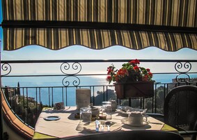 A table set for dining with a view of the sea, a potted plant with red flowers, and a sign indicating a no-smoking area.