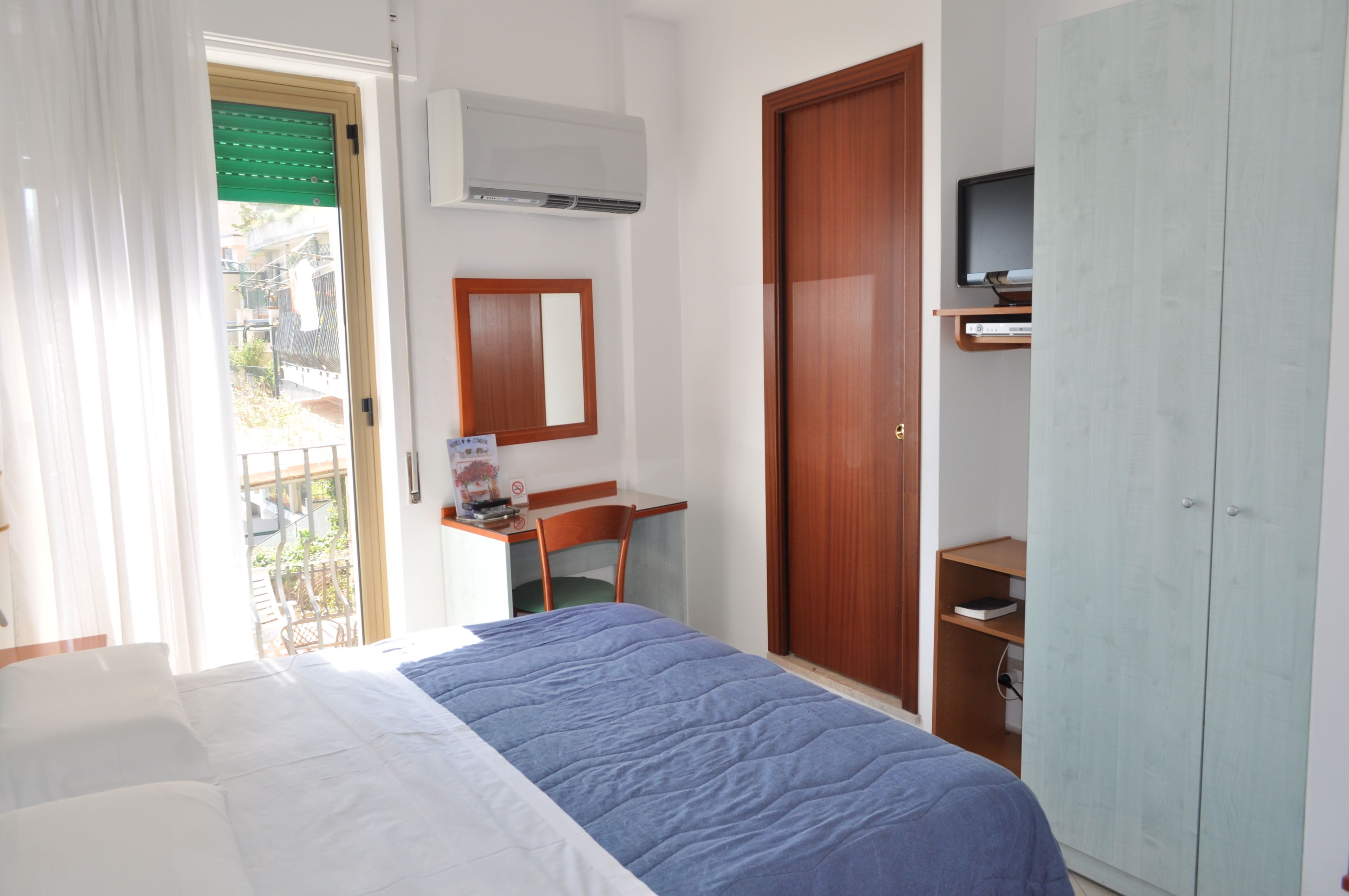 A bedroom with a bed, a desk, a mirror, a wardrobe, and an air conditioning unit.