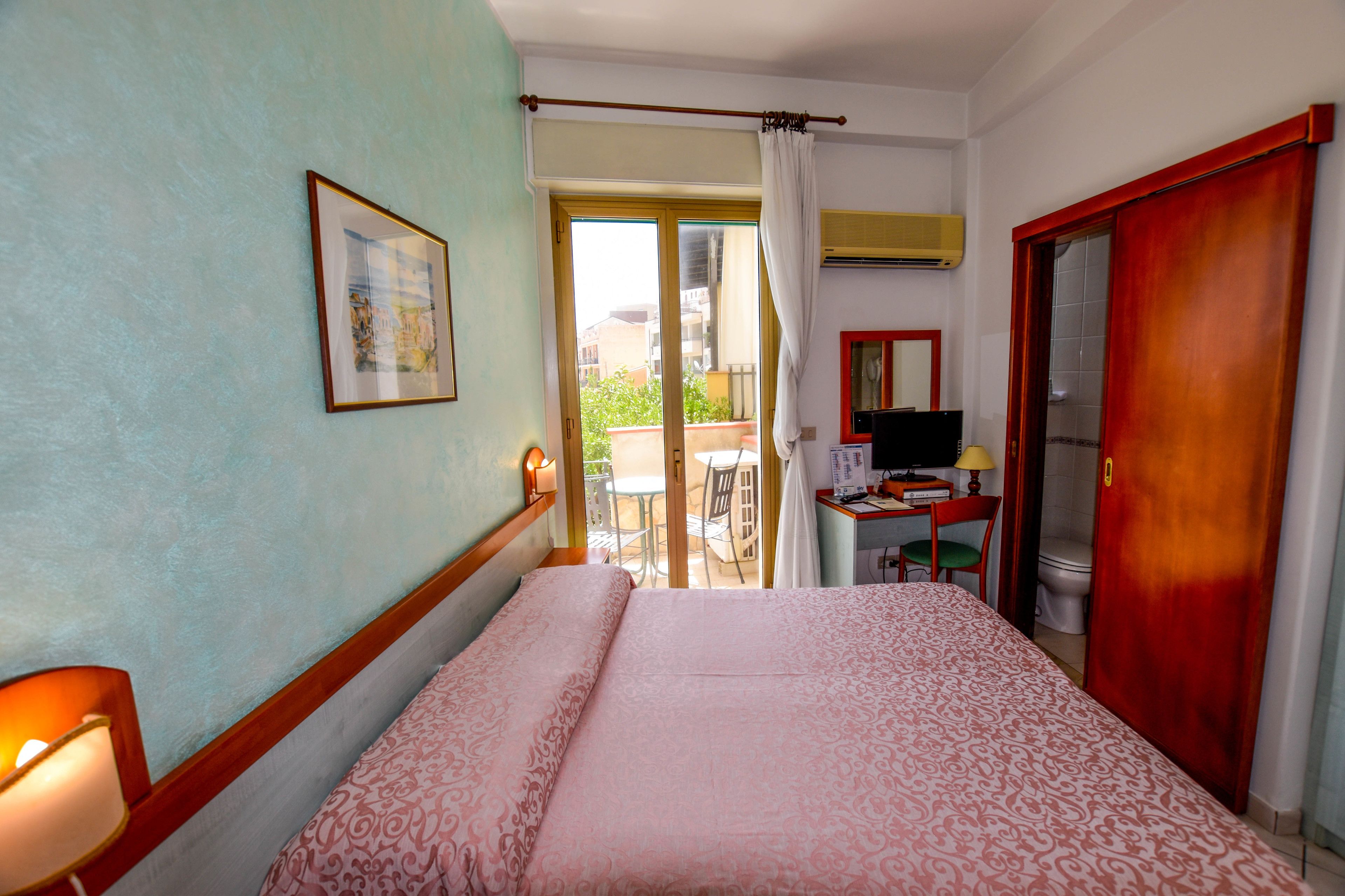 Habitación de hotel con cama, escritorio con silla, televisor y puerta corrediza a un área exterior.