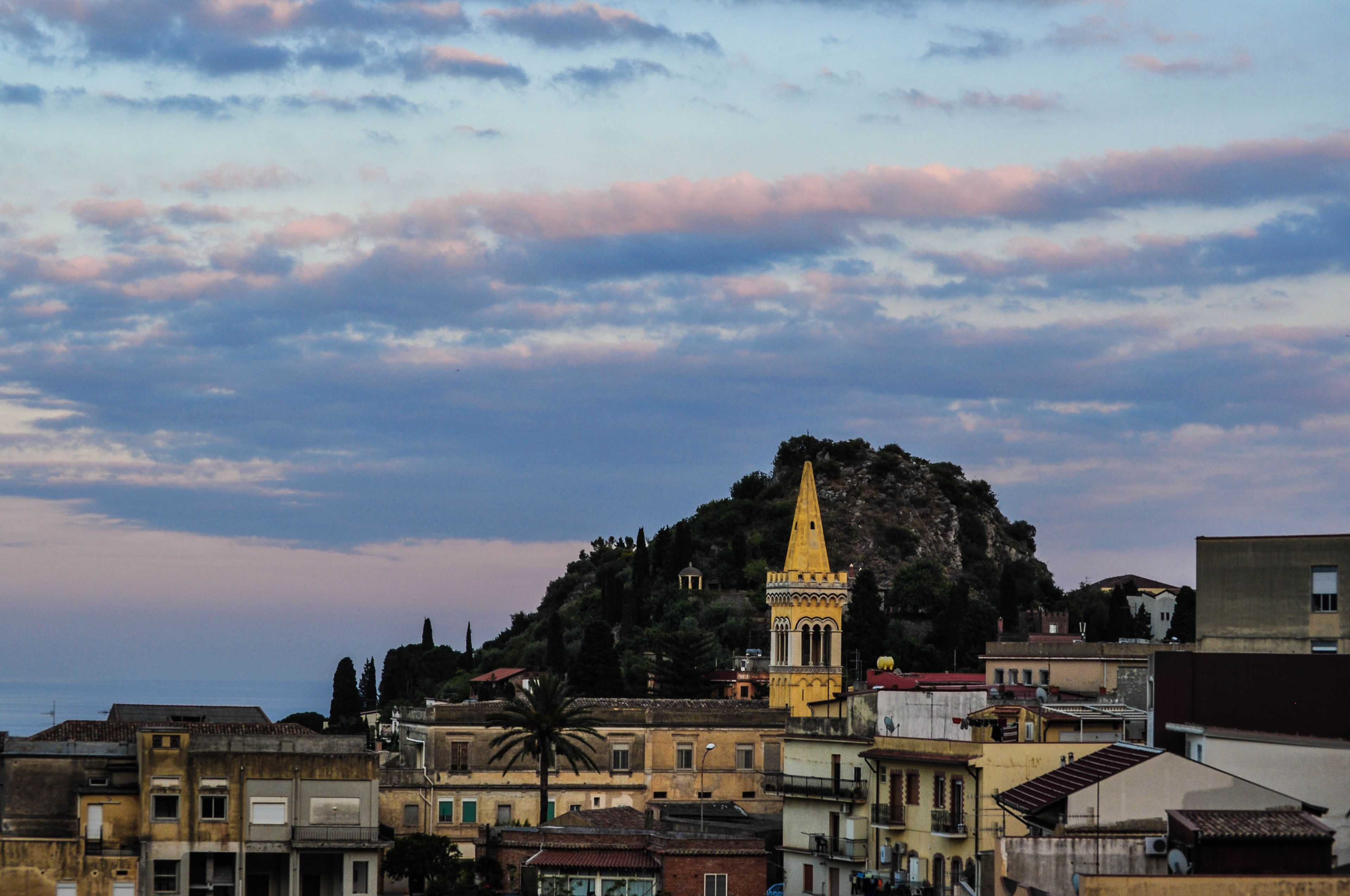 A coastal town with historic buildings, a prominent church with a tall bell tower, and a hill with dense vegetation.