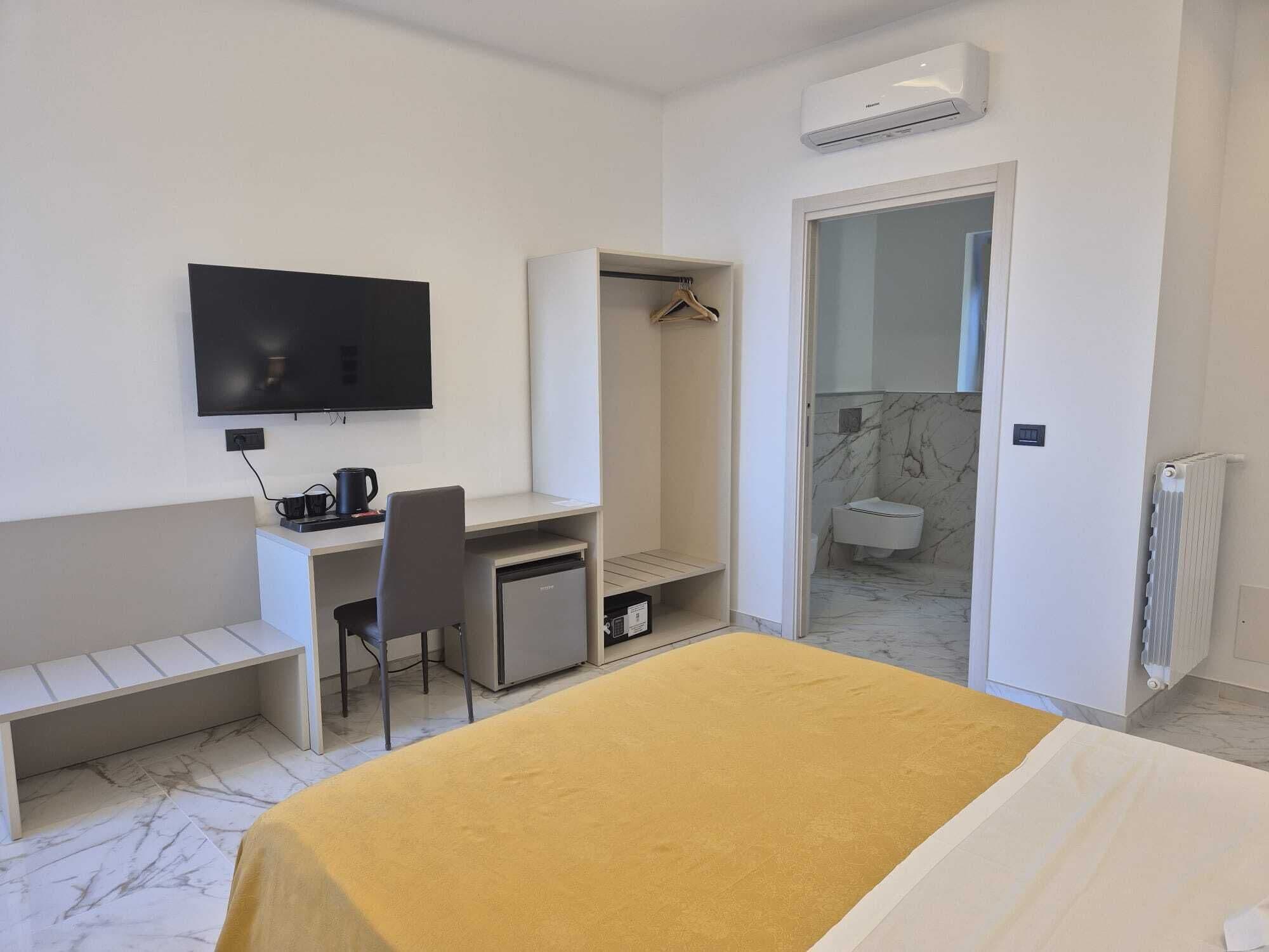 A hotel room with a bed, a desk with a chair, a TV, a wardrobe, and a bathroom visible through an open door.