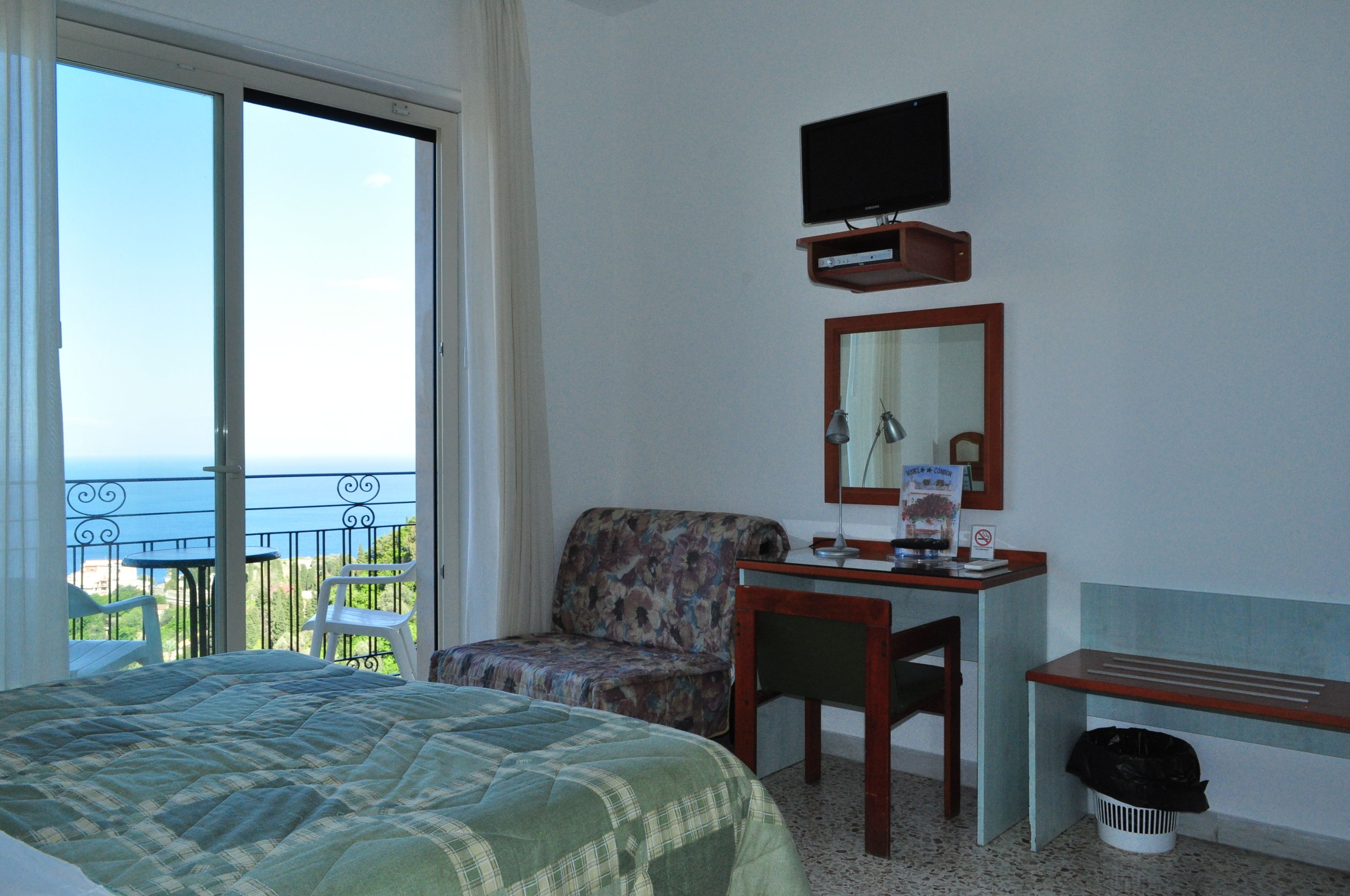 A hotel room with a bed, a desk, a chair, a television, and a view of the sea.